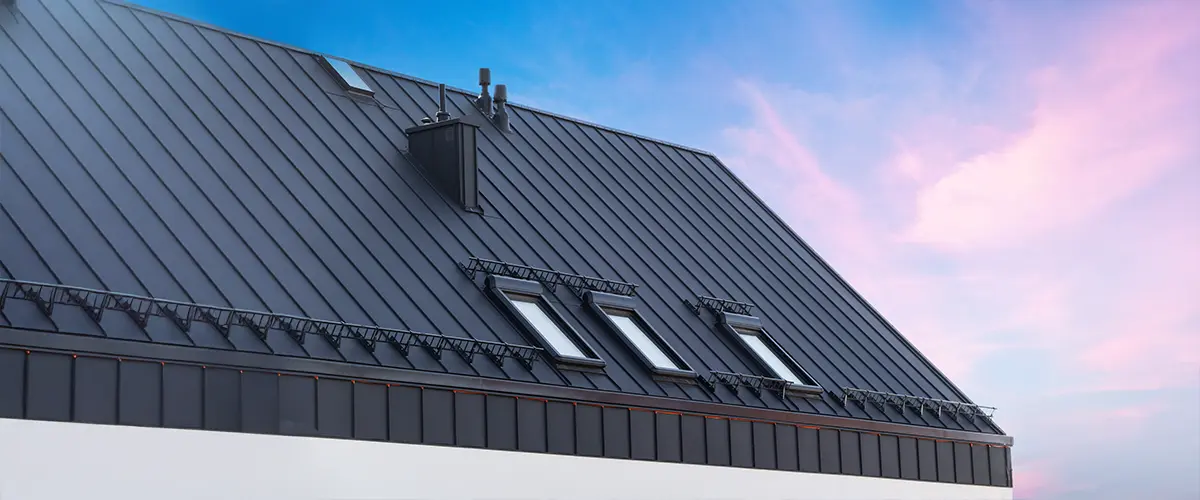 Modern black metal roof with skylights and chimney, set against a pink and blue sunset sky.