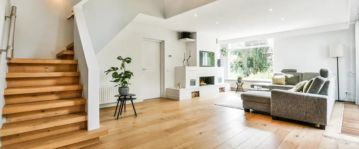 Modern living room with wooden staircase, hardwood flooring, gray sectional sofa, and ample natural light.