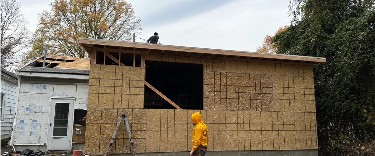 Residential home addition under construction with plywood walls, worker on roof, and scaffolding. New house extension project.