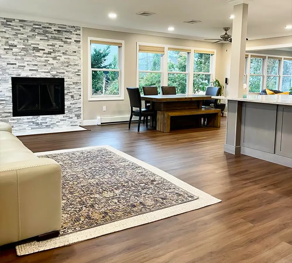 Spacious living room featuring a stone fireplace, hardwood floors, dining area, and large windows with natural light.