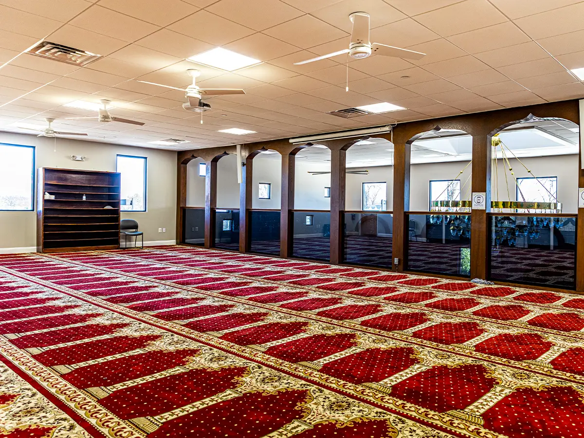 Spacious mosque prayer room with green carpets, ceiling fans, and a modern, open layout for worship.