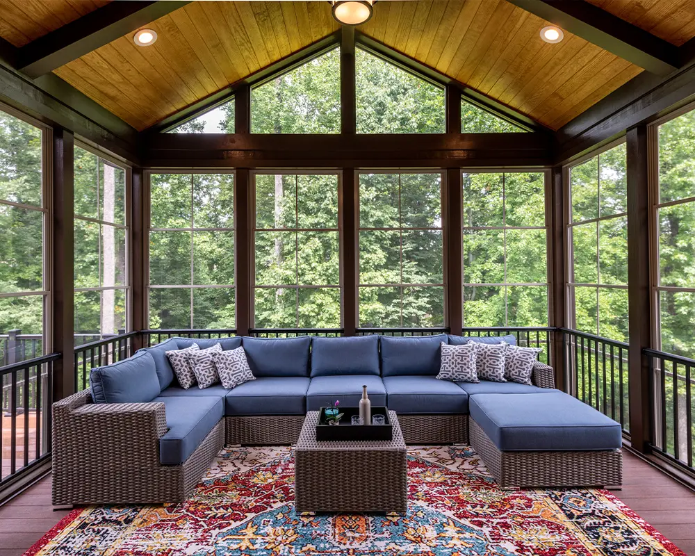 Cozy screened porch with blue sectional sofa, vibrant rug, and wooden ceiling, surrounded by lush trees.