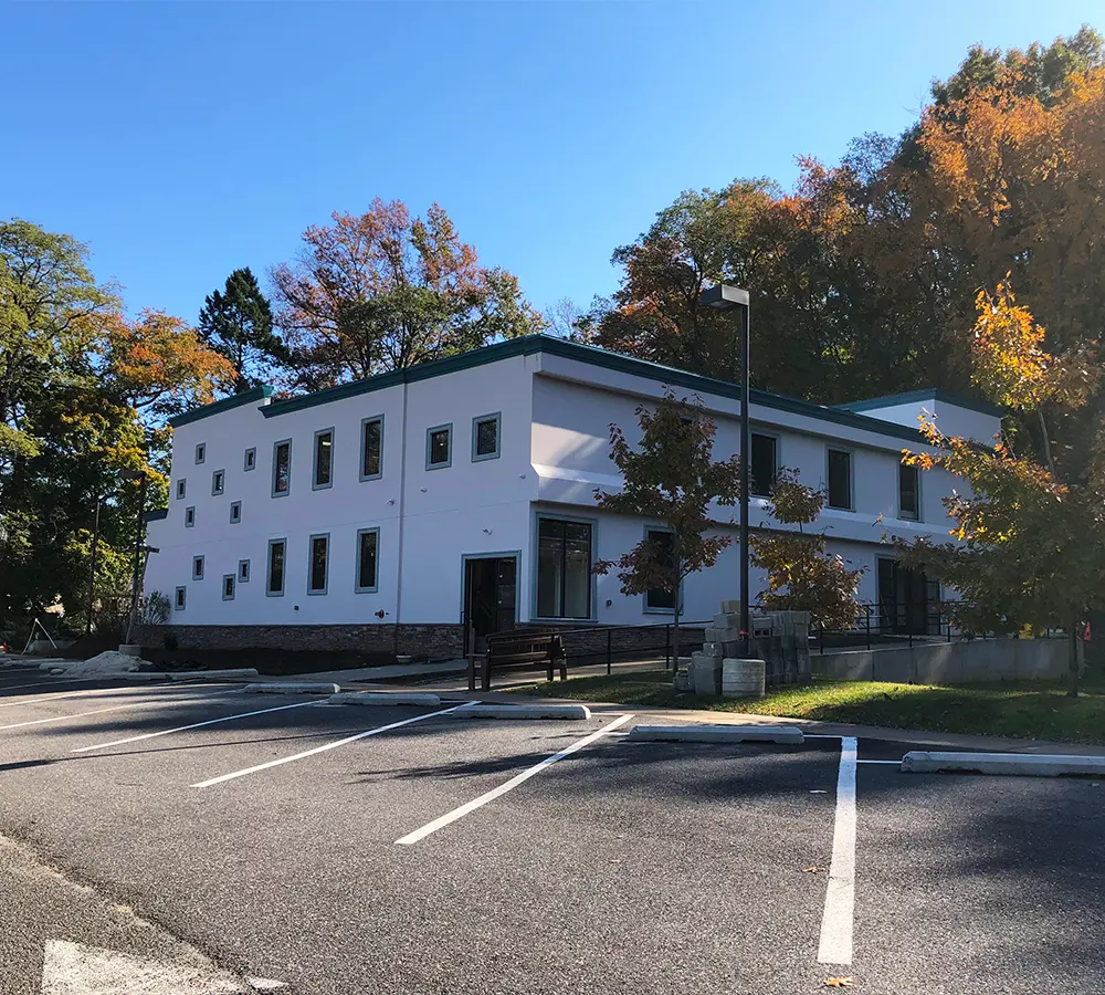 Contemporary two-story building featuring a white exterior, large windows, and a clean parking area surrounded by trees.