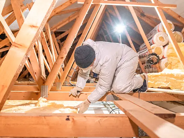 Worker installing attic insulation with fiberglass rolls in wooden-framed roof structure for energy efficiency.