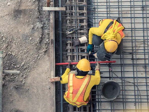 Construction workers in safety vests and helmets reinforcing steel rebar at a building foundation site.