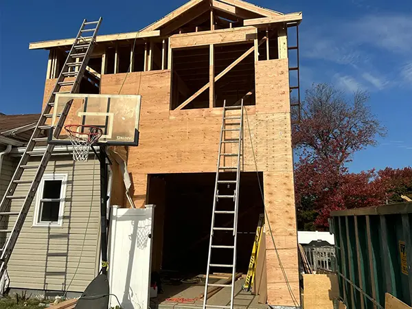 Two-story home addition under construction with wooden framing, ladders, and a basketball hoop in the driveway.