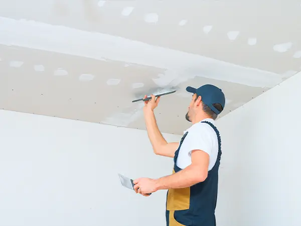 Construction worker applying joint compound to drywall ceiling during home renovation project with a trowel and scraper.