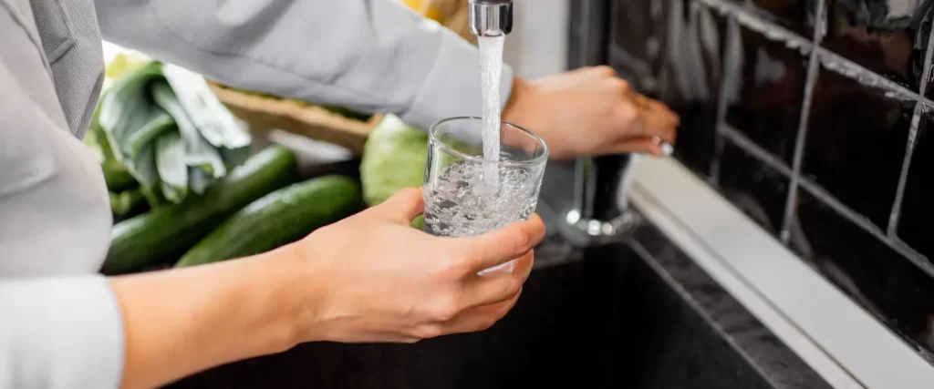 A person filling a glass with water from a kitchen faucet next to fresh vegetables.
