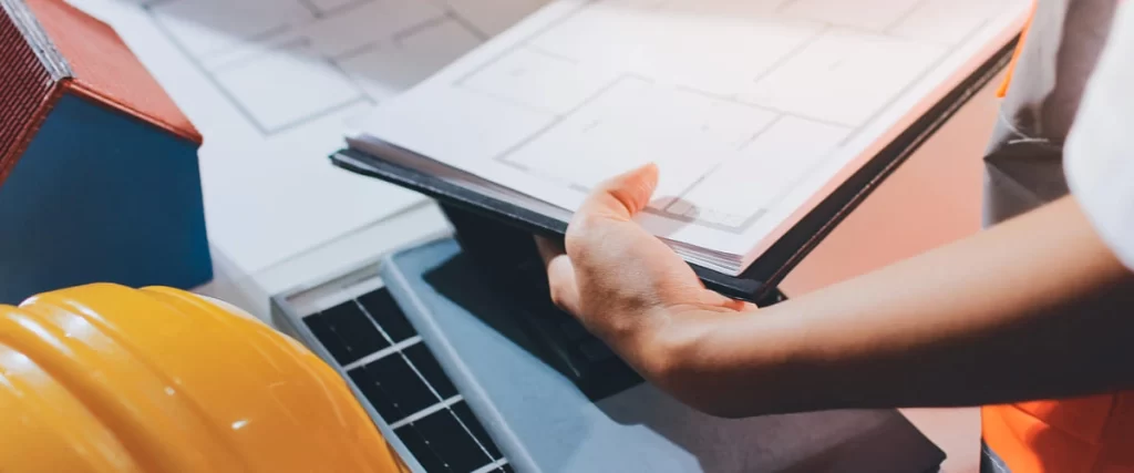 A construction professional organizing project documents on a desk next to a yellow safety helmet and a miniature model house.